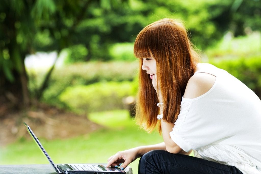 Young adult asian female sitting outdoors at a picnic table and reading her laptop computer.