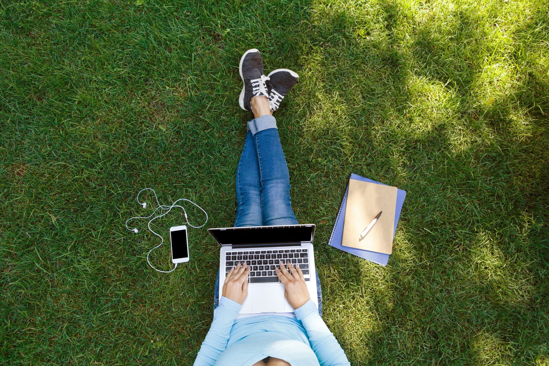 Top view of girl sitting in park on the green grass with laptop, notebook and smartphone with earphones, hands on keyboard.